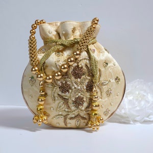 以下が含まれることがあります： クリーム色の巾着袋で、金色の刺繍とビーズのトリムが施されています。袋は丸い形をしていて、花柄で飾られています。袋の両側に金色のタッセルが付いています。