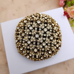Puede incluir: Un accesorio redondo negro cubierto de filigrana de metal dorado y brillantes pedrería transparente. La filigrana está dispuesta en un patrón floral, creando una superficie texturizada y ornamentada. Es probable que sea un artículo decorativo o una joya.
