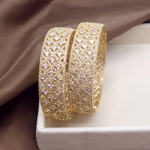 May include: Two gold-coloured bangles with a geometric pattern of square-cut, clear gemstones. The bangles are displayed on a white surface, with a brown fabric backdrop. The jewellery is designed with a repeating pattern of square shapes.
