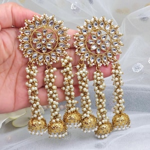 May include: Gold tone earrings with a round, floral design and multiple strands of white pearl beads. The earrings have a dangling bell charm at the bottom.