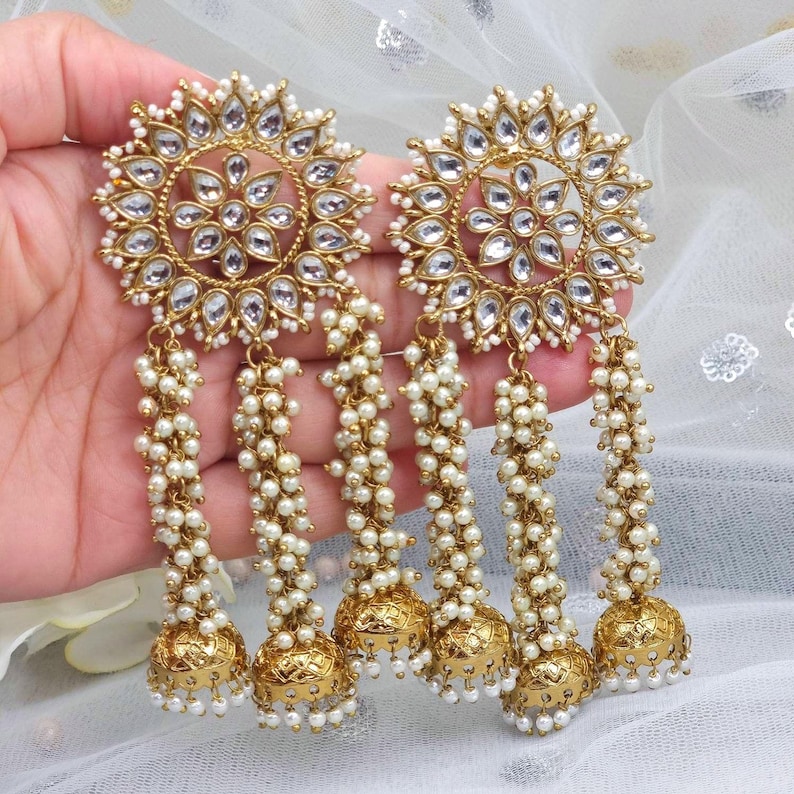 May include: Gold tone earrings with a round, floral design and multiple strands of white pearl beads. The earrings have a dangling bell charm at the bottom.