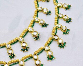 Può includere: Primo piano di gioielli color oro con pietre chiare a forma di goccia e piccole perline verdi. I gioielli presentano un design dettagliato con un mix di trame e forme, creando un aspetto elegante e ornato. Lo sfondo è un tessuto bianco morbido.