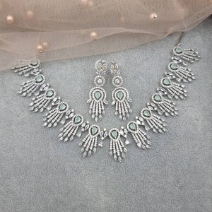May include: A silver-toned jewelry set featuring a necklace and matching earrings. The necklace and earrings have a repeating design of teardrop-shaped, light green stones surrounded by clear, sparkling crystals. The set is displayed on a gray surface.