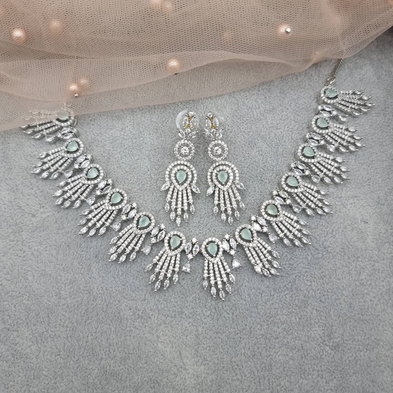 May include: A silver-toned jewelry set featuring a necklace and matching earrings. The necklace and earrings have a repeating design of teardrop-shaped, light green stones surrounded by clear, sparkling crystals. The set is displayed on a gray surface.
