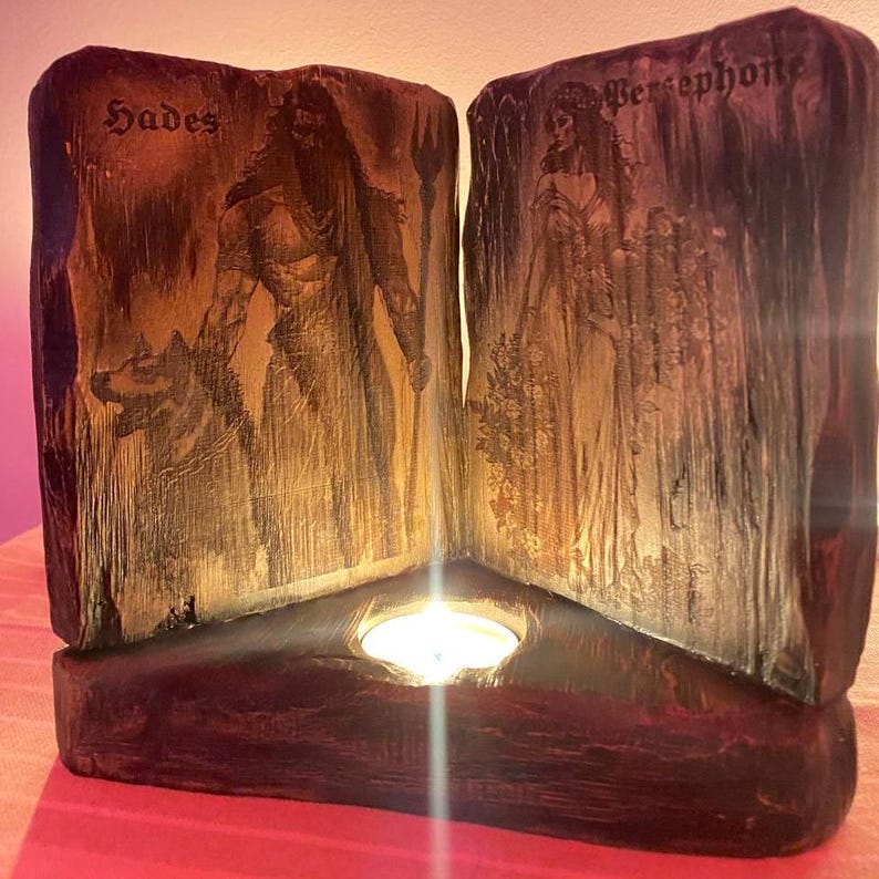 Może przedstawiać: Drewniany świecznik z tr&oacute;jkątną podstawą. Świecznik ma dwa panele z obrazami Hadesa i Persefony, z tekstem "Hades" i "Persephone" nad każdym obrazem. Zapalona świeca znajduje się w środku świecznika.