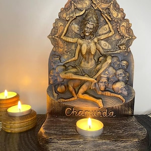May include: A carved wooden statue of Chamunda, a deity with multiple arms, seated on skulls, and set against an ornate backdrop. The word "Chamunda" is carved into the base. Lit candles in wooden holders are in the foreground.