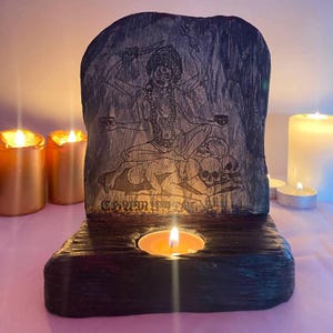 Può includere: Un portacandele in legno scuro intagliato con un'incisione dettagliata di una divinità con più braccia, seduta sopra teschi. Una piccola candela brucia nella base, illuminando l'opera d'arte. Ci sono candele aggiuntive sullo sfondo.