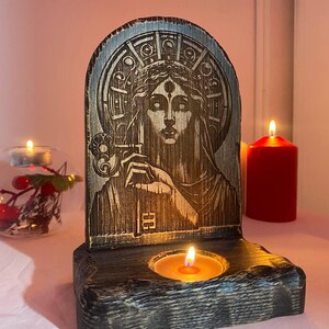 以下が含まれることがあります： 3つ目の女性が彫られた木製のキャンドルホルダー。ホルダーの中央には小さなキャンドルがあり、黒く塗られています。