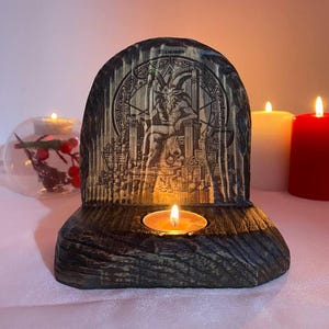 Puede incluir: Un portavelas de madera con una imagen tallada de una figura con cabeza de cabra sentada en un trono. El portavelas tiene una vela pequeña en el centro y está decorado con un acabado de madera oscura.