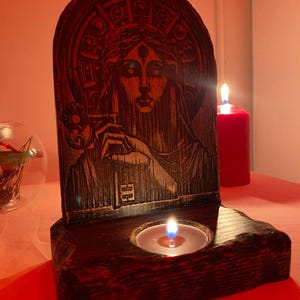 以下が含まれることがあります： 第三の目を描いた女性の彫刻が施された木製キャンドルホルダー。ホルダーの中央には小さなキャンドル、右には大きなキャンドルが置かれています。