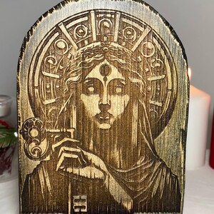 以下が含まれることがあります： 3つ目の目と鍵を持った女性の彫刻が施された木製キャンドルホルダー。女性は幾何学模様の円形デザインに囲まれています。キャンドルホルダーは濃い茶色に着色されています。