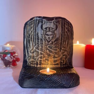 Könnte beinhalten: Ein Kerzenhalter aus Holz mit einer schwarzen und braunen Oberfläche. Der Halter zeigt eine geschnitzte Abbildung eines Dämons mit Flügeln und Hörnern. Der Text "Samari" ist auf dem Halter sichtbar. Eine kleine Kerze brennt im Halter.