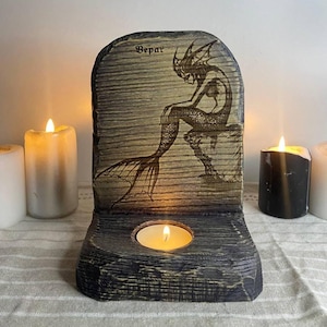 Puede incluir: Un portavelas de madera con un diseño de sirena tallada. La palabra "Bepar" está encima de la sirena. Una pequeña vela está encendida dentro del soporte. El soporte es gris y tiene un aspecto texturizado. El portavelas está rodeado de velas encendidas.