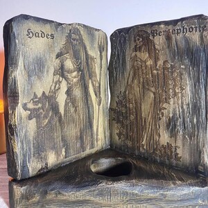 Może przedstawiać: Drewniana skrzynka z dwoma zawiasowymi panelami, każdy z czarno-białym obrazkiem. Lewy panel przedstawia Hadesa z wilkiem, a prawy panel przedstawia Persefonę. Skrzynka ma mały, okrągły otw&oacute;r w środku.