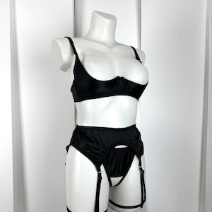 Op de afbeelding: Zwarte lingerie set op een witte mannequin. De set bevat een demi-cup bh met verstelbare bandjes en een hoge taille jarretelgordel met jarretels. De lingerie is gemaakt van een gladde, glanzende stof.