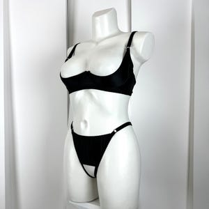 Op de afbeelding: Zwarte lingerie set op een witte mannequin. De set bevat een demi-cup bh met verstelbare bandjes en een bijpassende string met een uitgesneden ontwerp. De lingerie is gemaakt van een gladde, mogelijk satijnachtige stof.