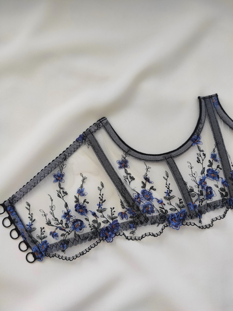 Peut inclure: Corset en dentelle noire avec broderie florale bleue. Le corset a un bord festonn&eacute; et est fait d'un tissu transparent.