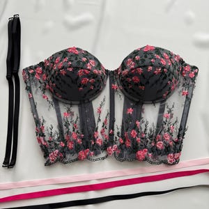 Peut inclure: Un corset en mesh noir avec des broderies florales roses et des bretelles r&eacute;glables. Le corset a une structure balein&eacute;e et une fermeture &agrave; busc &agrave; l'avant.