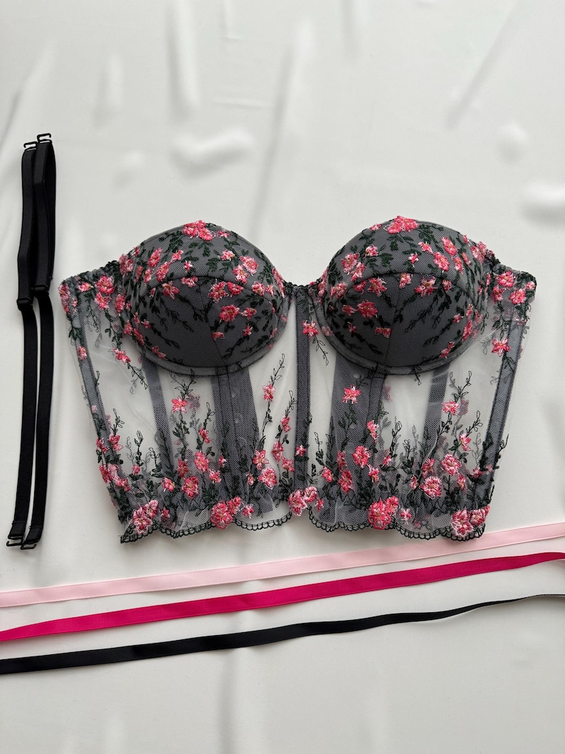 Peut inclure: Un corset en mesh noir avec des broderies florales roses et des bretelles r&eacute;glables. Le corset a une structure balein&eacute;e et une fermeture &agrave; busc &agrave; l'avant.
