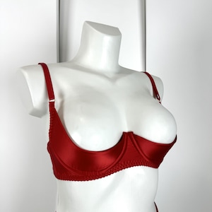 May include: A red satin bra with underwire support and adjustable straps. The bra is displayed on a white mannequin. The bra has a scalloped edge along the bottom and a classic, elegant design.
