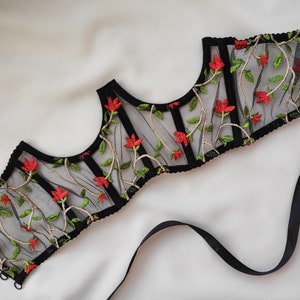 May include: Black mesh corset with red floral embroidery. The corset has a boned structure and is adorned with delicate floral patterns.