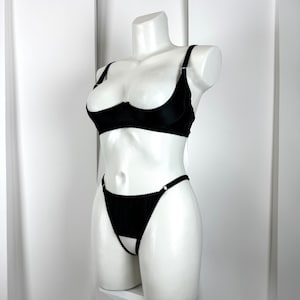 May include: Black lingerie set displayed on a white mannequin. The set includes a demi-cup bra with adjustable straps and a matching thong with a cut-out design. The bra and thong are made of a smooth, solid black fabric.
