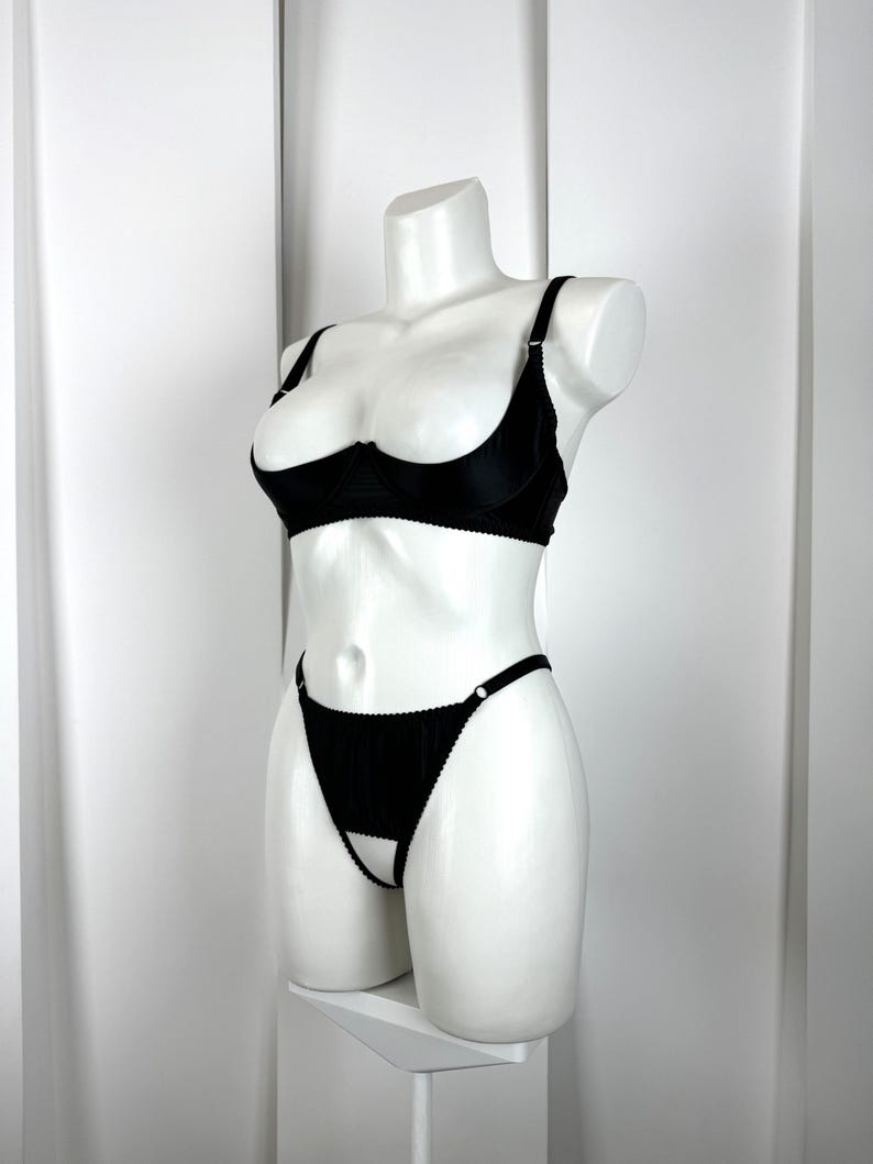 May include: Black lingerie set displayed on a white mannequin. The set includes a demi-cup bra with adjustable straps and a matching thong with a cut-out design. The bra and thong are made of a smooth, solid black fabric.