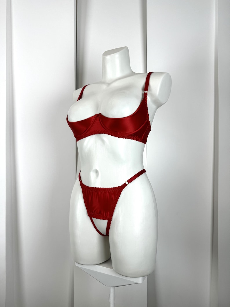 May include: A red lingerie set displayed on a white mannequin. The set includes a demi-cup bra and a thong. The bra has adjustable straps and the thong has a cut-out design. The fabric has a satin sheen.