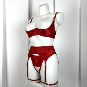 Op de afbeelding: Een rode lingerie set op een witte mannequin. De set bestaat uit een beha, jarretelgordel en string. De beha heeft verstelbare bandjes. De jarretelgordel heeft jarretelbandjes met goudkleurige metalen sluitingen. De lingerie is gemaakt van glanzend satijn.