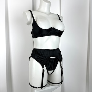 May include: Black lingerie set displayed on a white mannequin. The set includes a demi-cup bra with adjustable straps and a high-waisted garter belt with suspender straps. The lingerie is made of a smooth, shiny fabric.