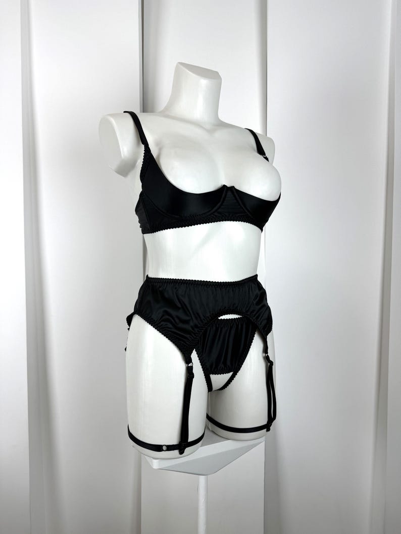May include: Black lingerie set displayed on a white mannequin. The set includes a demi-cup bra with adjustable straps and a high-waisted garter belt with suspender straps. The lingerie is made of a smooth, shiny fabric.