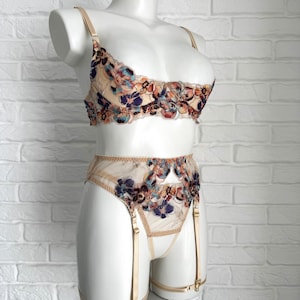 Op de afbeelding: Een beige lingerie set met bloemenborduurwerk in tinten oranje, bruin, blauw en paars. De set bestaat uit een bh en een jarretelgordel met verstelbare banden.