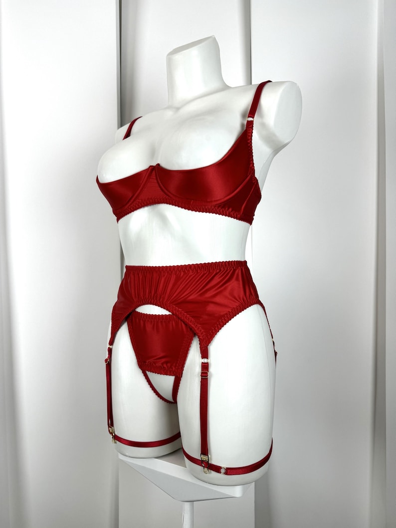 May include: A red satin lingerie set displayed on a white mannequin. The set includes a bra, garter belt, and matching underwear. The bra has adjustable straps and underwire support. The garter belt features adjustable straps and gold-colored hardware.