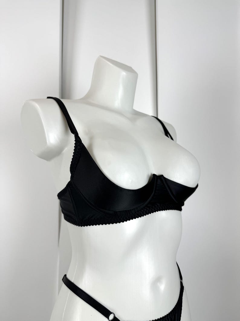 May include: A black bra and matching underwear set displayed on a white mannequin. The bra features underwire support and delicate trim along the edges. The lingerie set is made of a smooth, shiny fabric.