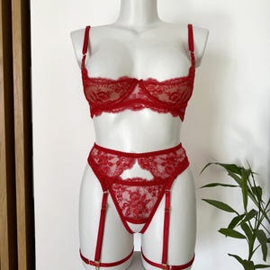 May include: A red lingerie set displayed on a mannequin. The set includes a bra with floral lace detailing, a matching high-waisted panty, and garter straps. The bra has adjustable straps. The lingerie is made of a sheer, delicate fabric.