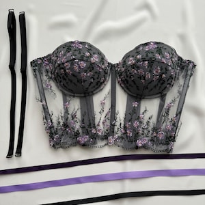 Peut inclure: Un corset noir en dentelle transparente avec des broderies florales violettes. Le corset est dot&eacute; d'une structure &agrave; baleines et d'un ourlet festonn&eacute;. Il est livr&eacute; avec deux bretelles noires et trois rubans violets.
