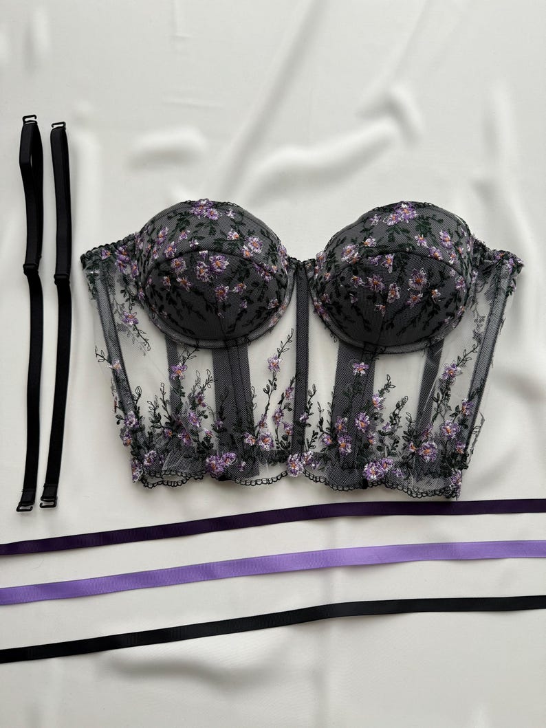 Peut inclure: Un corset noir en dentelle transparente avec des broderies florales violettes. Le corset est dot&eacute; d'une structure &agrave; baleines et d'un ourlet festonn&eacute;. Il est livr&eacute; avec deux bretelles noires et trois rubans violets.