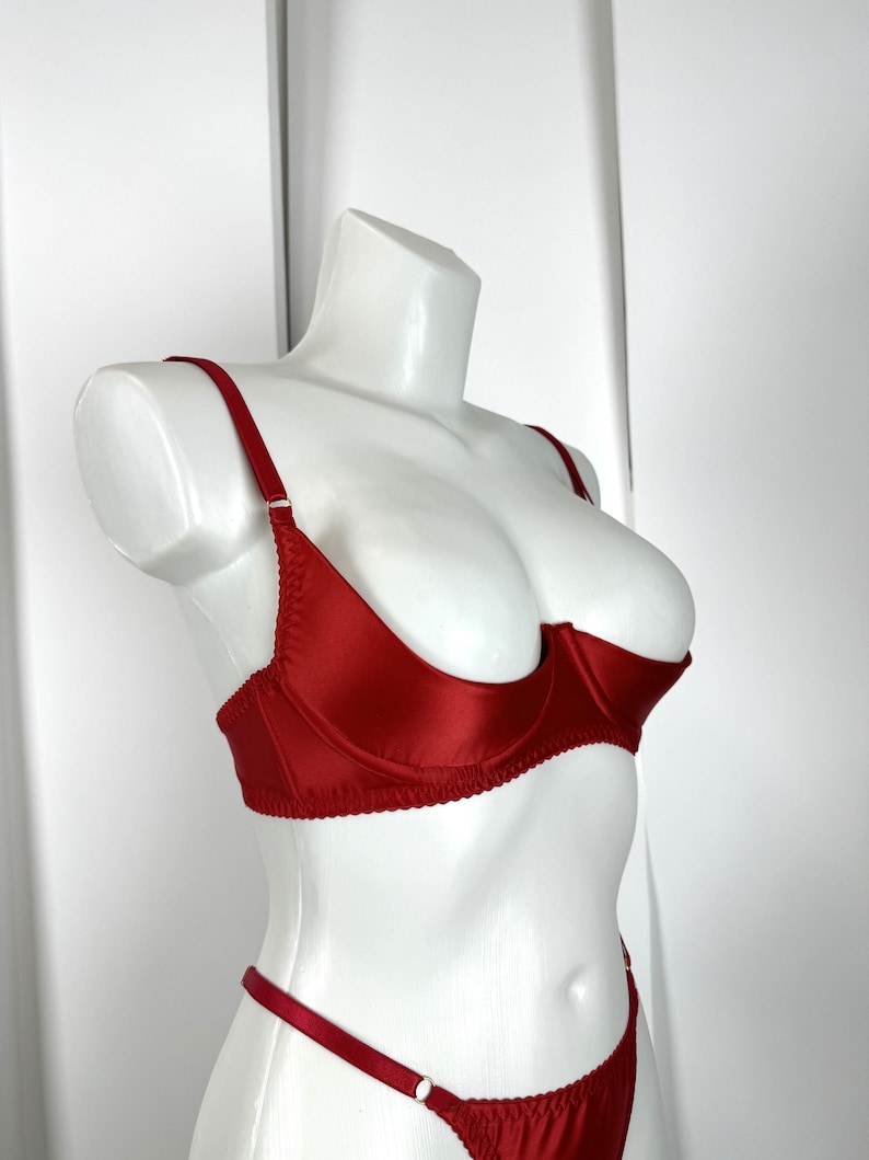 May include: A red satin lingerie set displayed on a white mannequin. The set includes a balconette bra with adjustable straps and a matching thong. The bra features underwire support and delicate stitching. The lingerie is a vibrant shade of red.