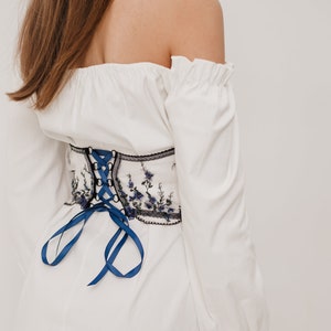 Peut inclure: Un haut blanc &agrave; &eacute;paules d&eacute;nud&eacute;es avec des manches longues et un ruban bleu nou&eacute; dans le dos. Le haut est dot&eacute; d'une ceinture corset brod&eacute;e de fleurs noires et blanches.