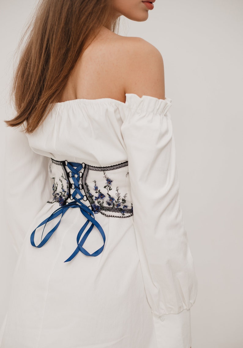 Peut inclure: Un haut blanc &agrave; &eacute;paules d&eacute;nud&eacute;es avec des manches longues et un ruban bleu nou&eacute; dans le dos. Le haut est dot&eacute; d'une ceinture corset brod&eacute;e de fleurs noires et blanches.