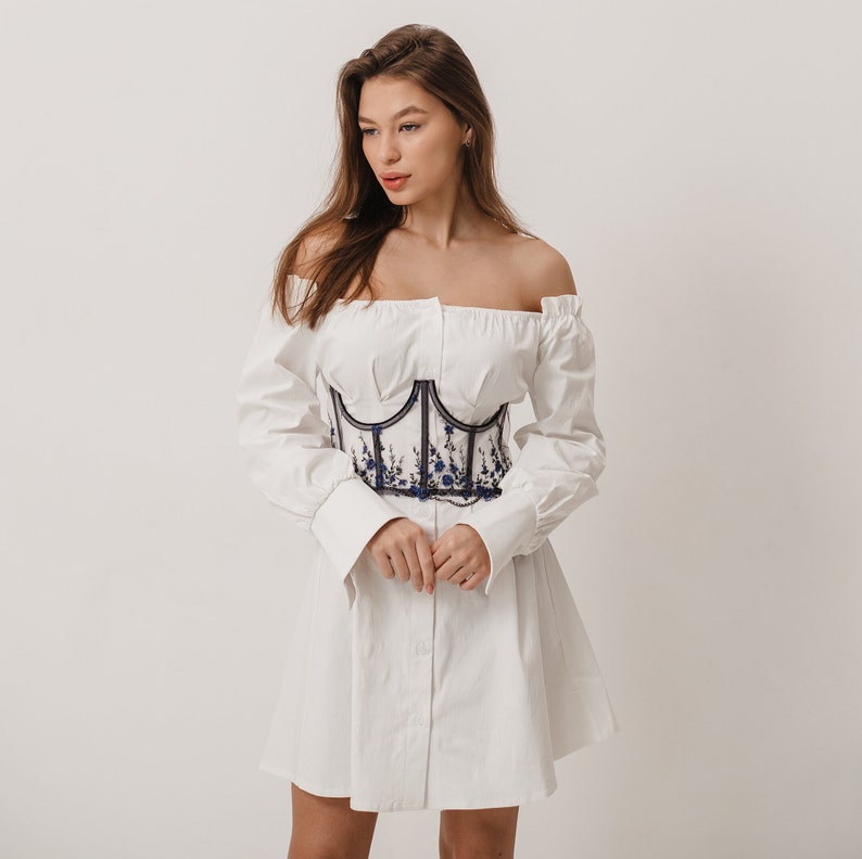 Peut inclure: Une robe blanche &agrave; &eacute;paules d&eacute;nud&eacute;es avec des manches longues et une ceinture corset brod&eacute;e de fleurs noires et bleues.