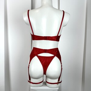 May include: A red lingerie set displayed on a mannequin. The set includes a bra, garter belt, and thong. The bra has thin straps and a back closure. The garter belt features adjustable straps and metal clasps. The thong has a high-cut design.