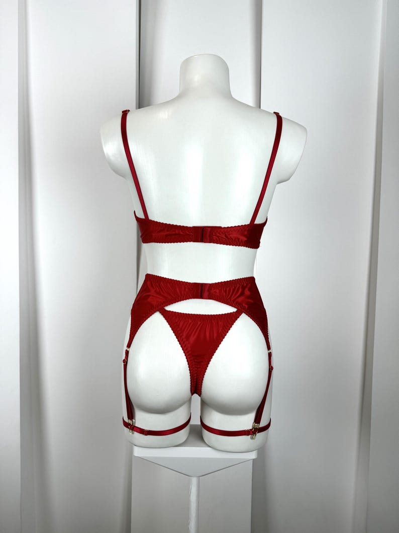 May include: A red lingerie set displayed on a mannequin. The set includes a bra, garter belt, and thong. The bra has thin straps and a back closure. The garter belt features adjustable straps and metal clasps. The thong has a high-cut design.