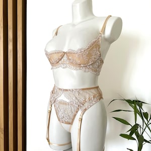 Op de afbeelding: Een beige lingerieset op een mannequin. De set bestaat uit een bh, een slip met hoge taille en een jarretelgordel. De bh en slip zijn versierd met delicate kanten details. De jarretelgordel heeft verstelbare bandjes.