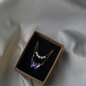 Pu&ograve; includere: Una collana con catena dorata con un ciondolo a forma di farfalla viola e una catena pi&ugrave; piccola con quattro perle bianche. La collana &egrave; in una scatola marrone con interno nero.