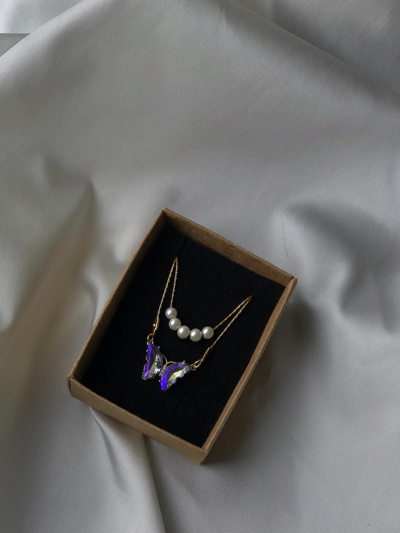Pu&ograve; includere: Una collana con catena dorata con un ciondolo a forma di farfalla viola e una catena pi&ugrave; piccola con quattro perle bianche. La collana &egrave; in una scatola marrone con interno nero.