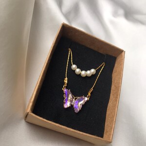Pu&ograve; includere: Una delicata collana a catena dorata con un ciondolo a forma di farfalla iridescente viola e una fila di perle bianche. La collana &egrave; esposta in una scatola marrone con interno nero.