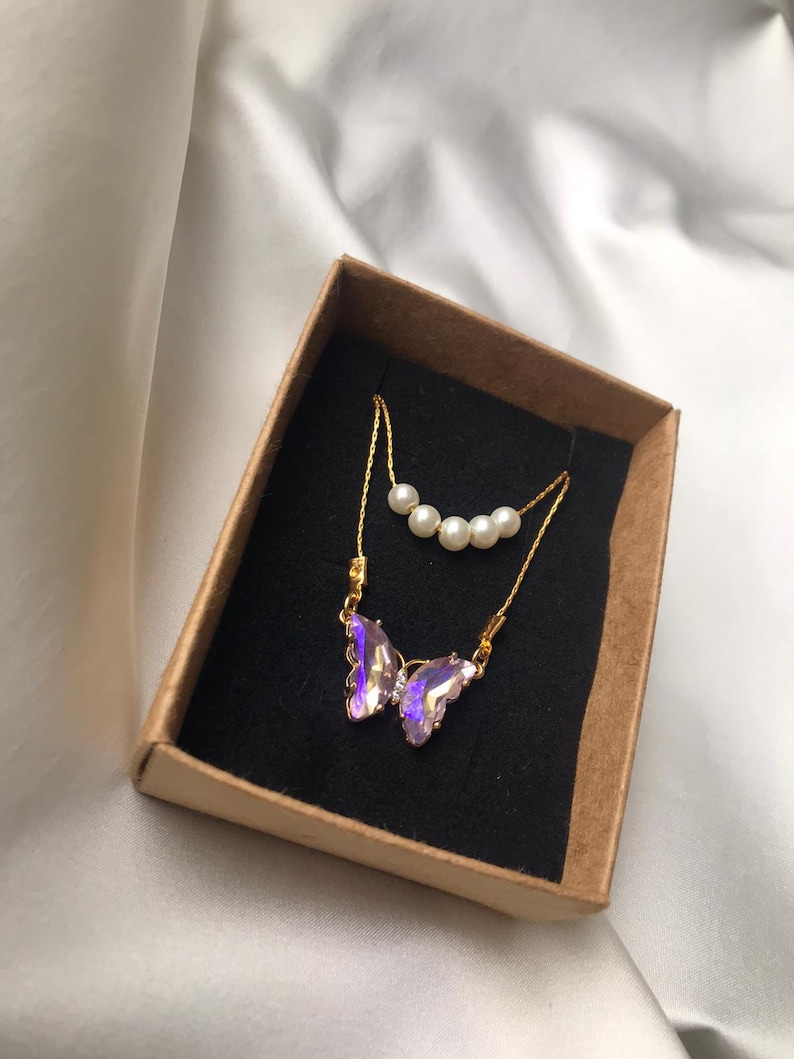 Pu&ograve; includere: Una delicata collana a catena dorata con un ciondolo a forma di farfalla iridescente viola e una fila di perle bianche. La collana &egrave; esposta in una scatola marrone con interno nero.