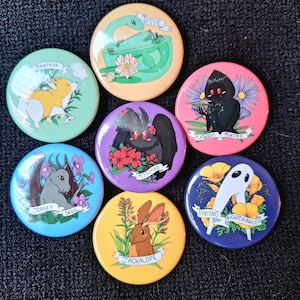 May include: Set of ten 2.25 inch diameter round button pins featuring various cryptids and mythical creatures. Each pin has a different creature, name, and location. The pins are colorful and feature a variety of floral designs.