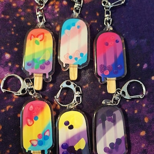 May include: Six colorful popsicle keychains with different fruit designs. Each keychain features a popsicle with a wooden stick and a silver key ring. The popsicles are decorated with various fruits, including strawberries, blueberries, and lemons.